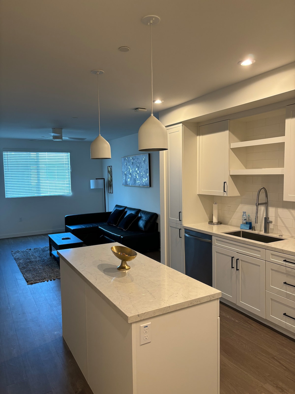A modern kitchen area features sleek white cabinets and stainless-steel appliances. An island with a countertop offers space for dining or preparation. A cozy living area is visible, with a dark sofa and a decorative rug enhancing the overall contemporary design.