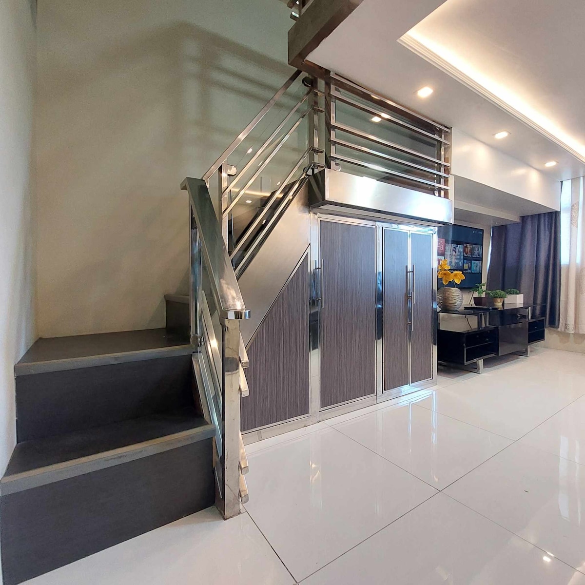 A modern stairway with sleek metal railings leads to an upper level. The landing features cabinetry with a wood finish, complemented by a vibrant arrangement of yellow flowers. Bright white tiles reflect natural light from a nearby window, enhancing the space's contemporary design.