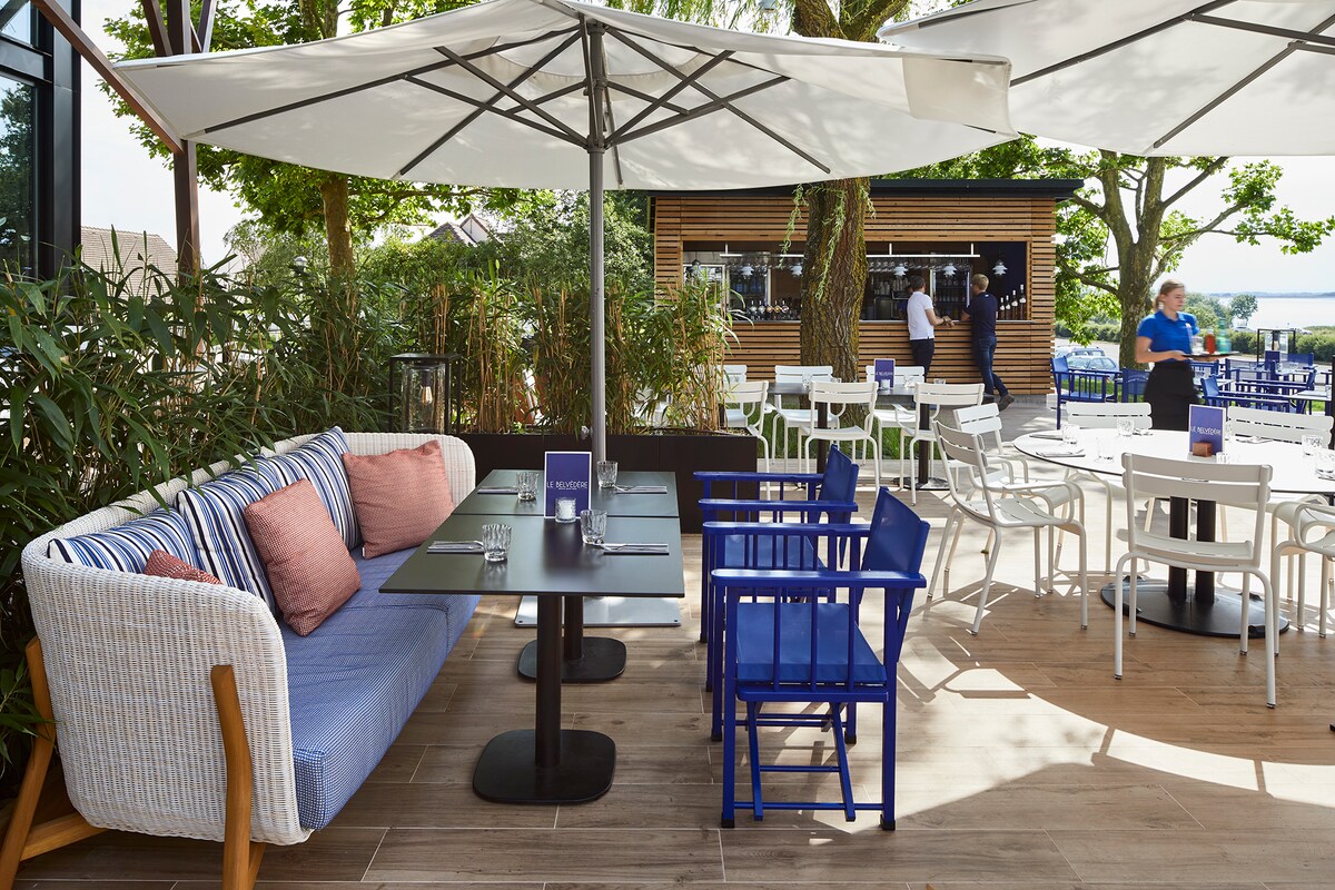 An outdoor dining area features numerous tables and chairs under large umbrellas, providing shade. A mix of colorful blue and white furniture creates a casual atmosphere. Lush greenery surrounds the space, with a bar visible in the background.
