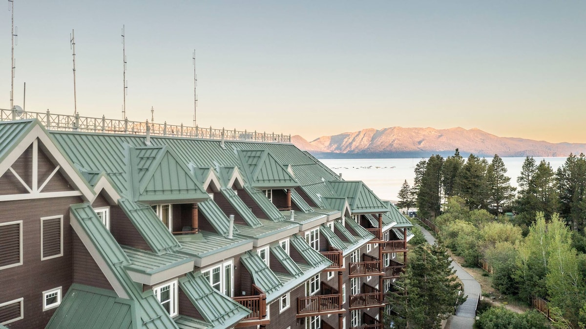 The image captures the exterior of a multi-story resort building with a green metal roof. Surrounding trees and mountains are visible in the background, highlighting a serene natural setting. The area suggests spacious balconies attached to the units, enhancing the inviting atmosphere.
