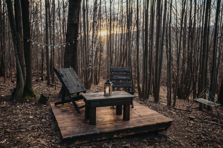 A wooden platform is set in a forest clearing, featuring two chairs and a table. A lantern sits atop the table, surrounded by tall trees. The backdrop includes a sunset visible through the branches, creating a serene outdoor setting.
