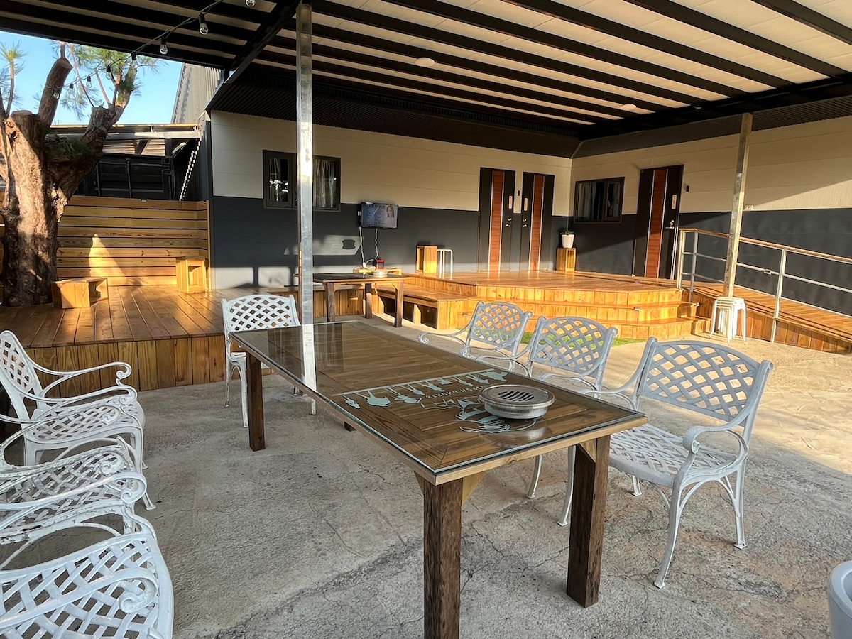 An outdoor gathering space is featured, with a large wooden table surrounded by white metal chairs. The area showcases a wooden deck with steps leading to cozy seating options. Accommodations are visible in the background with sliding doors and a television mounted on the wall.