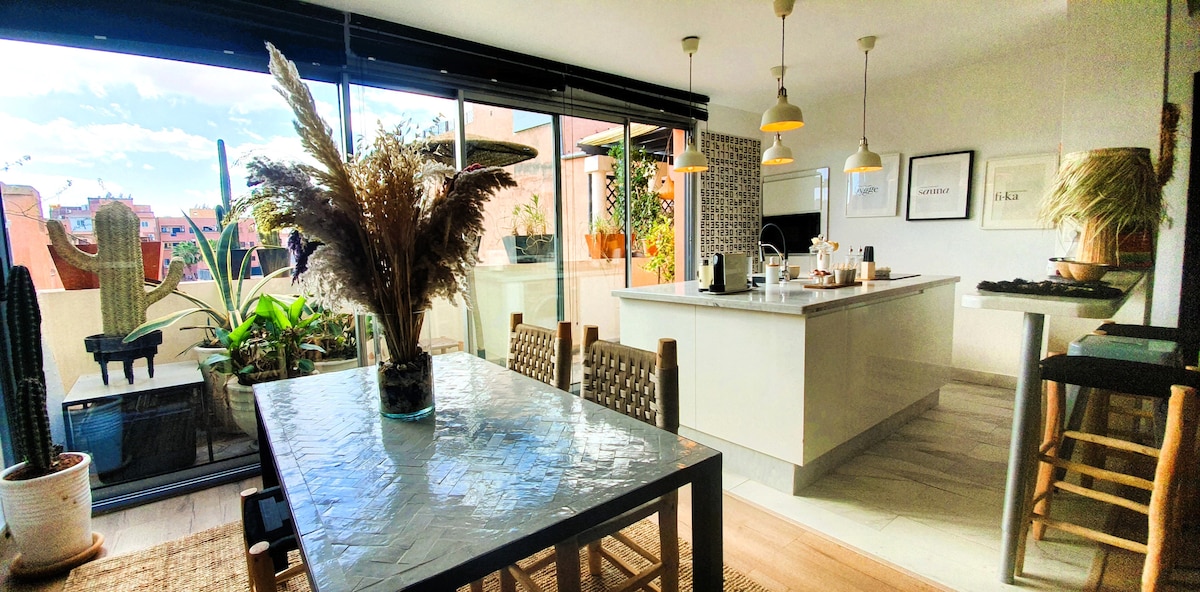 A modern dining area features a large table with decorative plants nearby. Bright light floods the space through expansive glass doors, revealing a well-equipped kitchen with a central island. Chic bar stools complement the elegant decor, creating an inviting atmosphere for meals and gatherings.