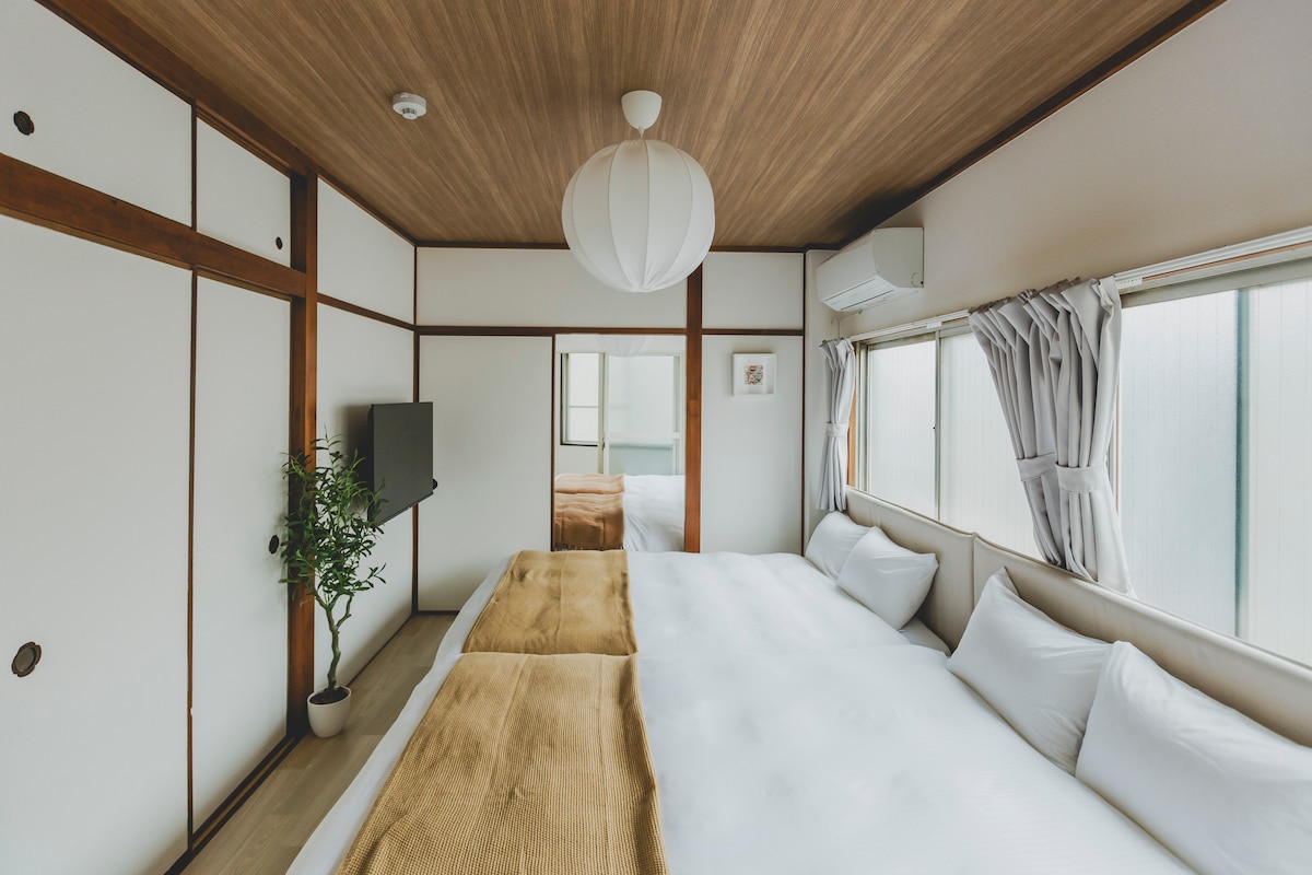 A well-lit bedroom features two spacious beds with soft white linens. A large window allows natural light to fill the room, complemented by light-colored curtains. A decorative plant stands in the corner, and a wall-mounted television is visible.