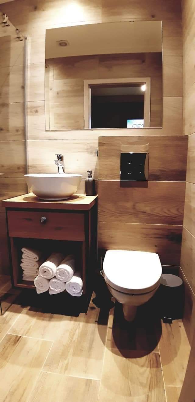 The bathroom features wood-toned walls and flooring, creating a natural ambiance. A modern sink is situated on a wooden vanity, accompanied by neatly arranged rolled towels. A mirror hangs above the sink, while a compact toilet completes the space.
