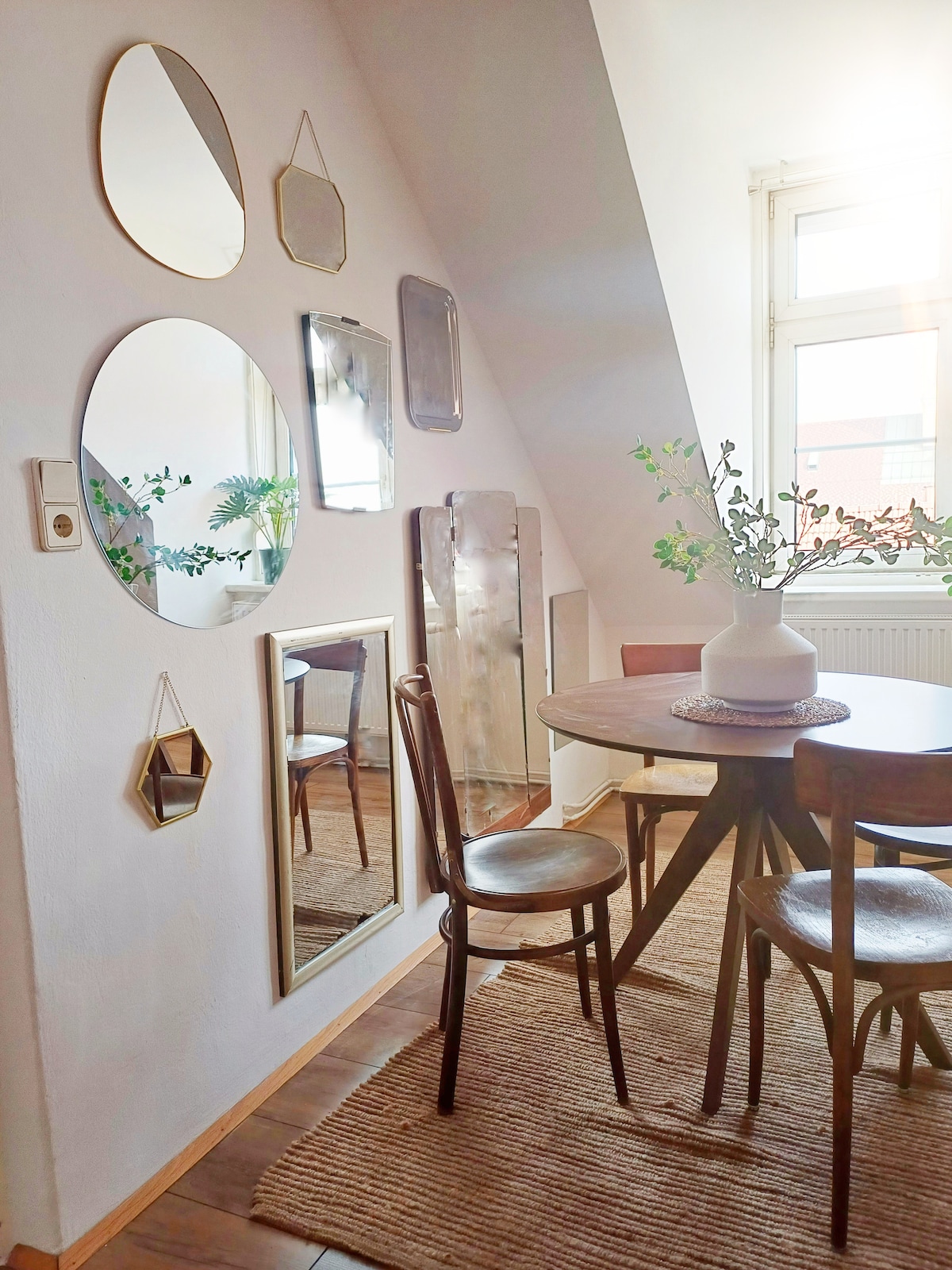 A bright dining area features a round table surrounded by wooden chairs. Mirrors of various shapes adorn the wall, reflecting light from the nearby window. A neutral-toned rug adds warmth to the space, while a vase with greenery sits elegantly on the table.