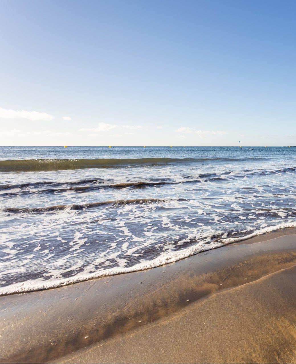 A serene beach scene is seen, featuring gentle waves lapping against a light brown sandy shore. The water displays varying shades of blue, reflecting a clear sky, which offers a calm and inviting atmosphere.