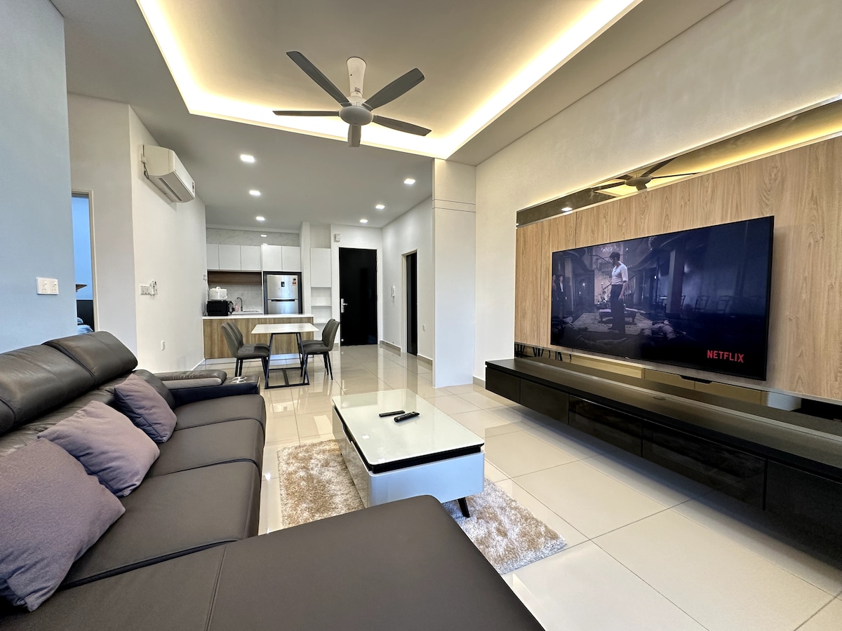 A modern living space features a comfortable sectional sofa adorned with decorative cushions. A large screen TV is mounted on the wall opposite a sleek coffee table. The open layout connects to the dining area and kitchen, highlighted by contemporary furnishings and ample natural light.