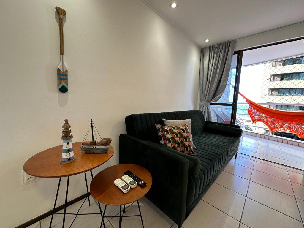 A modern living area features a dark green sofa adorned with a patterned cushion. Two round wooden tables display remote controls and decorative items. Natural light enters through large glass doors, leading to a balcony with a vibrant hammock.