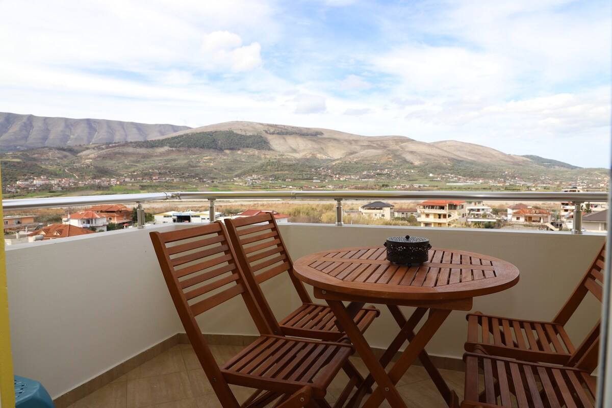 A balcony area is showcased with a round wooden table surrounded by four wooden chairs. The view includes green hills and distant mountains, contributing to a serene outdoor space.
