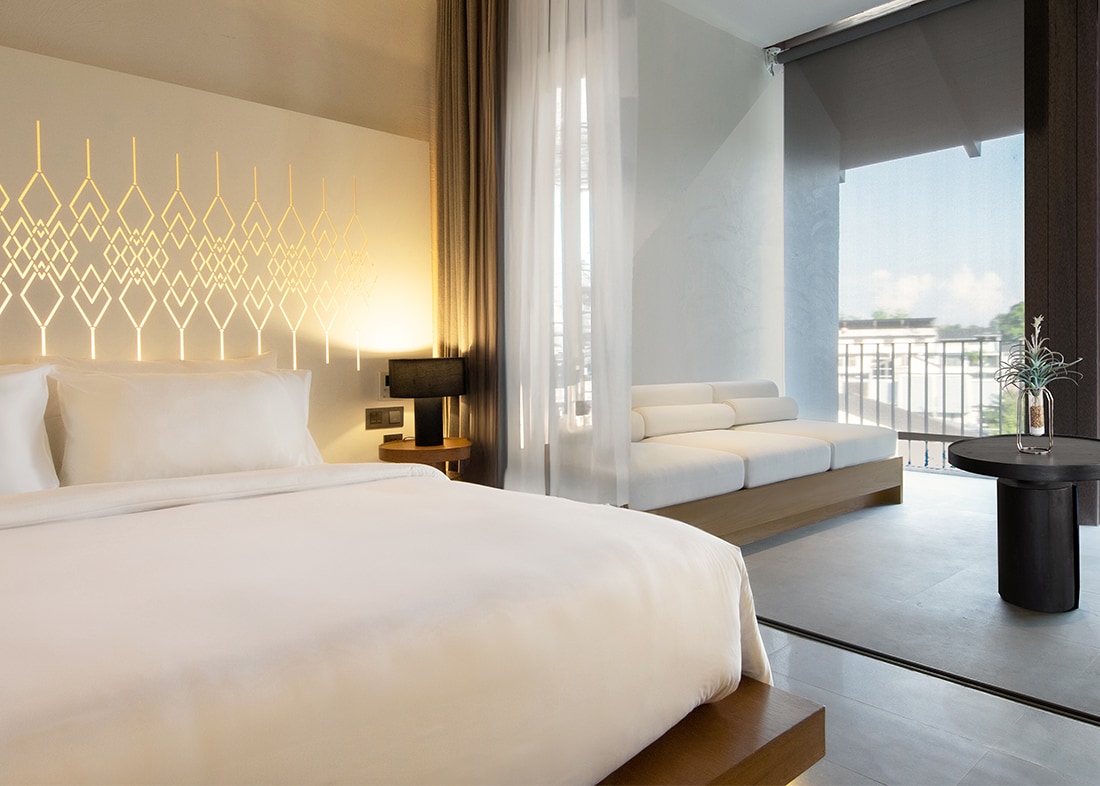 A spacious room features a large bed with soft linens, complemented by a stylish accent wall with an elegant design. Natural light fills the space through floor-to-ceiling windows, while a cozy seating area on the balcony offers a serene view.