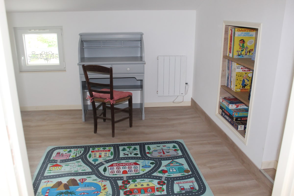 A cozy room features a wooden chair with a pink cushion positioned near a grey desk. A colorful area rug depicting a town layout is placed on the floor. A small bookshelf is visible, holding various board games, beside a window that allows natural light to enter.