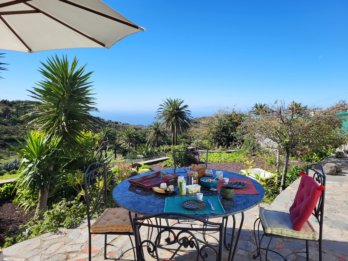 An outdoor dining area is set with a round table surrounded by leafy palm trees. The table displays a colorful spread of food and drinks. A bright blue sky and distant ocean views create a serene backdrop.