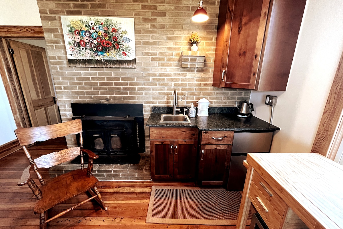 A functional kitchenette features a brick accent wall and wooden cabinetry. A kitchen sink is positioned alongside a black countertop with essential appliances, including a kettle. A rustic wooden chair is placed nearby, complementing the warm tones of the wood flooring.
