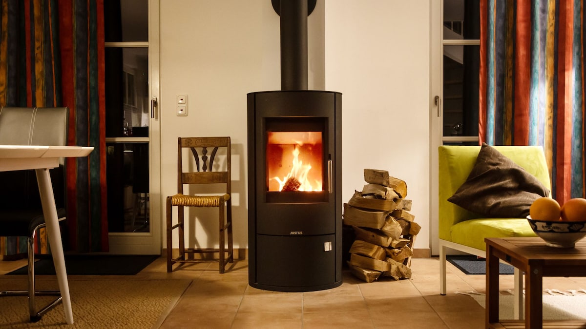 A modern wood stove is centrally placed, with a visible fire providing warmth. Surrounding it are a stack of firewood and a vibrant green armchair. Colorful curtains frame a glass door, while a dining table and wooden chairs create a functional and inviting space.