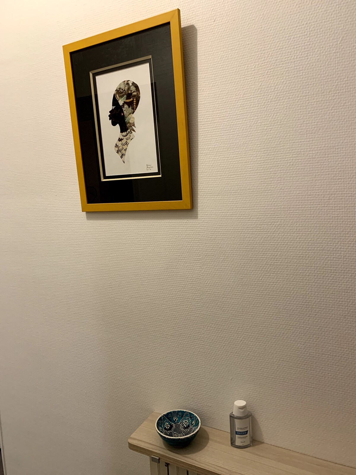 A framed artwork with a silhouette design is displayed on the wall, complemented by a yellow frame. Below, a small decorative bowl sits on a light-colored surface, with a bottle positioned nearby, enhancing the minimalist decor of the space.