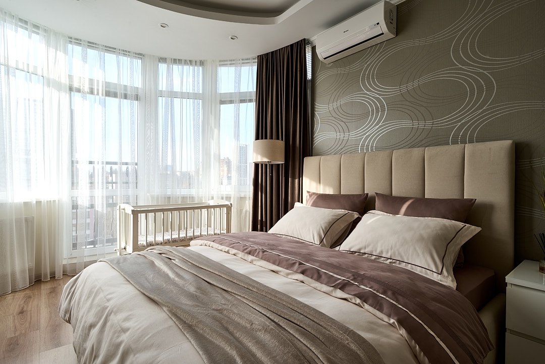A well-lit bedroom features large windows draped with sheer white curtains, allowing ample natural light. The spacious bed is adorned with neutral-colored linens, complemented by decorative pillows. A modern lamp and a minimalistic nightstand are placed beside the bed, enhancing the room's serene atmosphere.