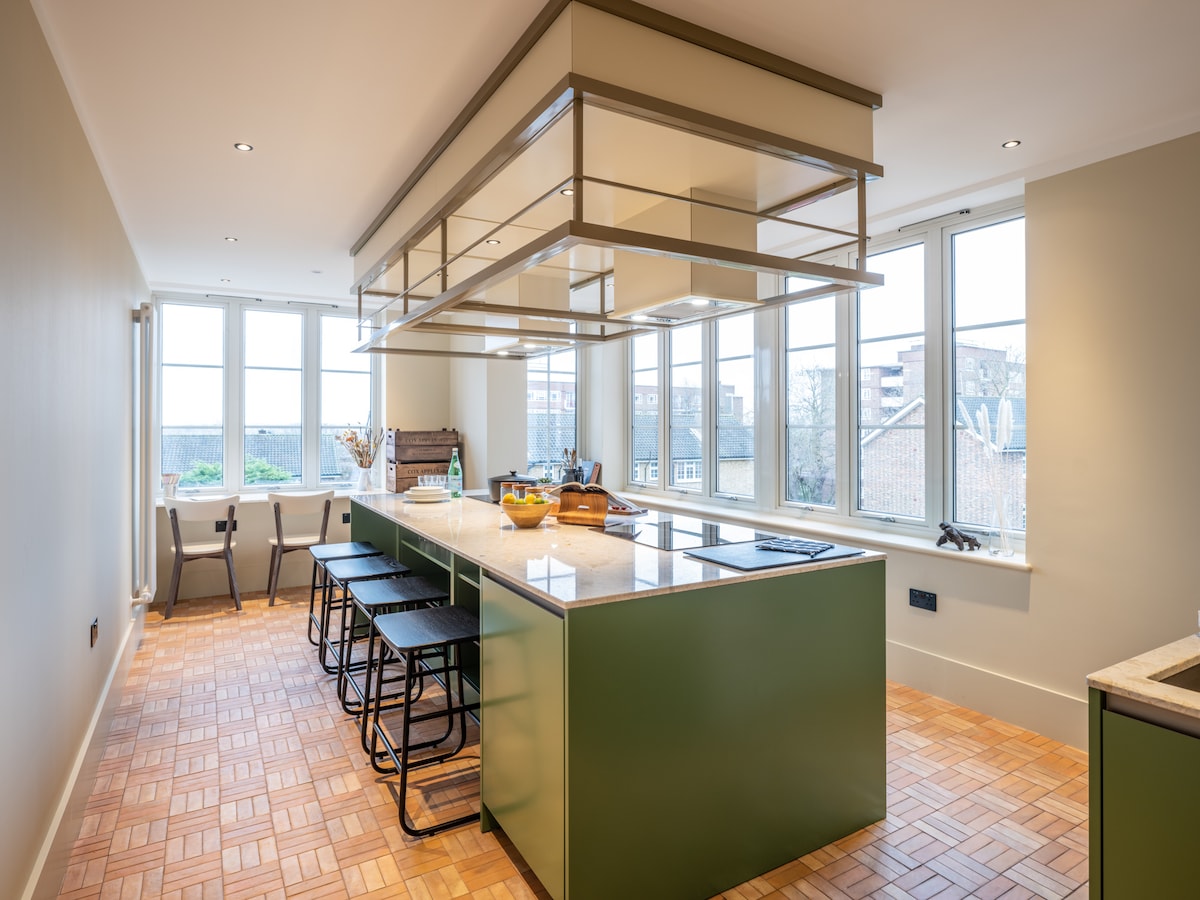 A modern kitchen is featured with a spacious island countertop and bar seating for four. Large windows provide ample natural light, highlighting the green cabinetry and light wood flooring. A decorative arrangement is placed on the island, adding a touch of warmth to the contemporary design.