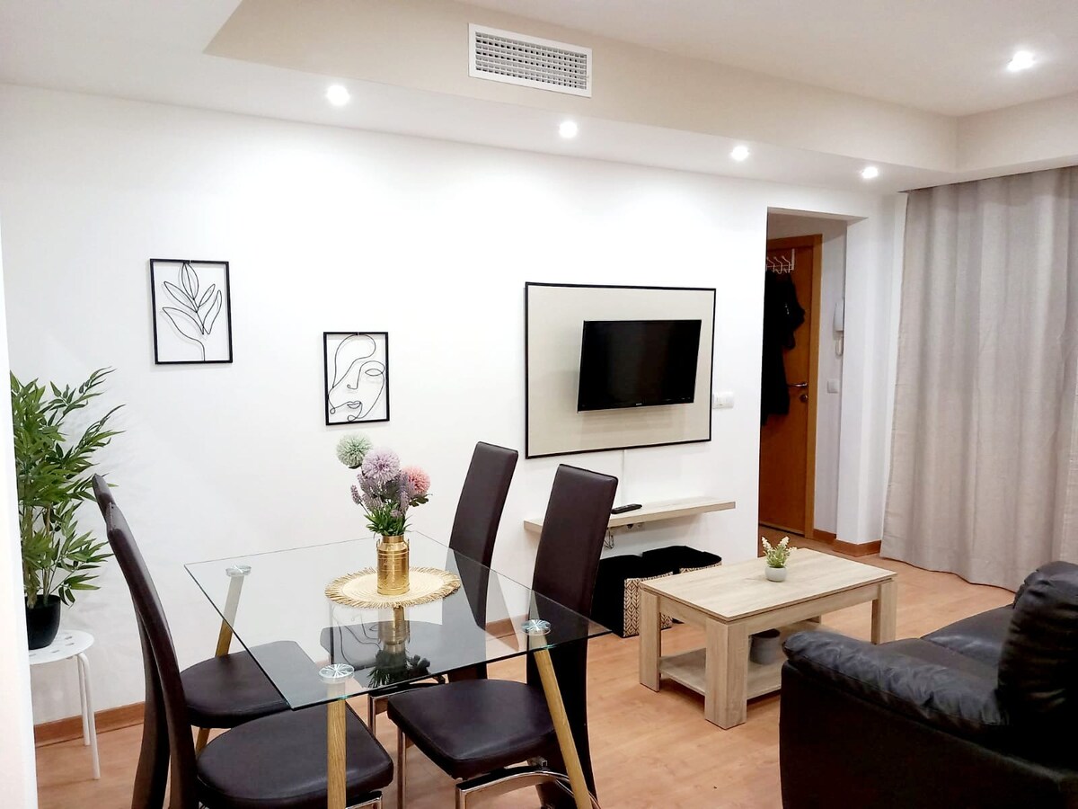 A modern dining area features a glass table surrounded by four dark chairs, with framed botanical art on the walls. A light wooden coffee table sits adjacent to a plush black sofa, providing a comfortable seating arrangement. A TV is mounted on the wall across from the sofa.