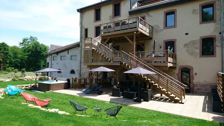 A spacious outdoor area features wooden decks leading up to the building, with shaded seating under patio umbrellas. Lounge chairs and a hot tub are arranged on a grassy lawn, surrounded by mature trees and a serene atmosphere.