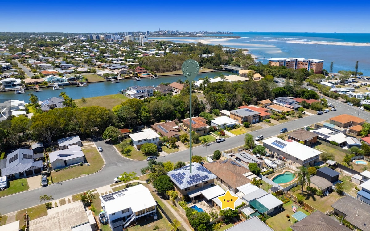 An aerial view captures the coastal neighborhood of Golden Beach, showcasing a mix of residential homes with well-maintained gardens. The surrounding waterways and nearby beach are visible, along with a glimpse of the Sunshine Coast skyline in the distance.