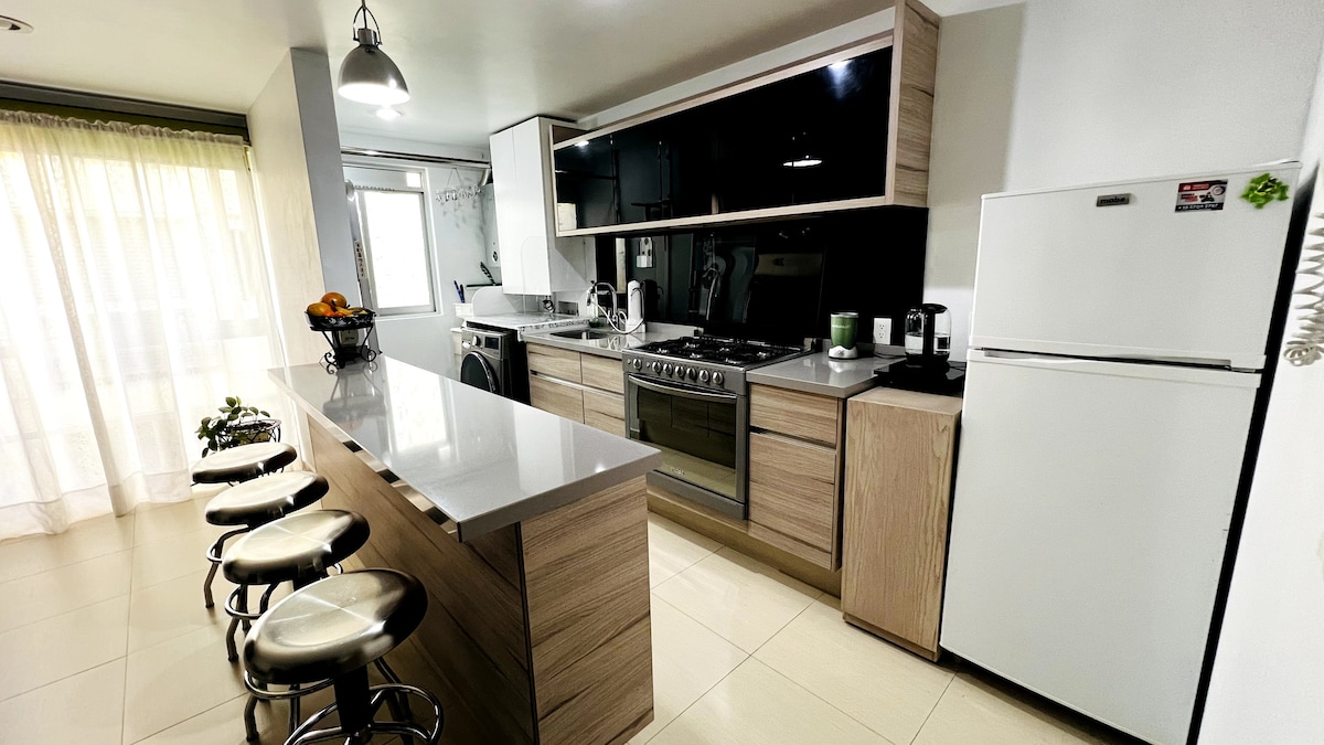 A modern kitchen features light wood cabinetry and sleek countertops. Stainless steel appliances, including a gas stove and refrigerator, are visible. Four bar stools are arranged around a central island, while a large window allows natural light to fill the space.