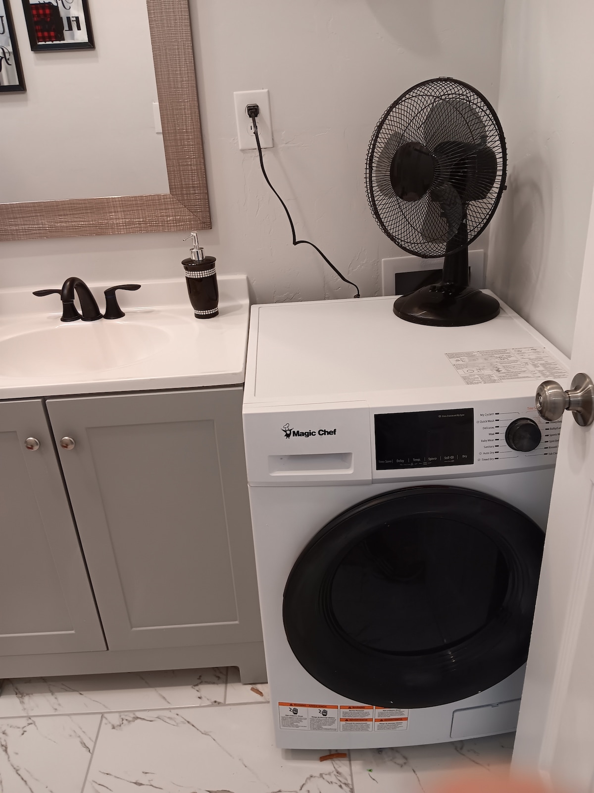 A compact washer and dryer unit is positioned next to a bathroom sink with modern fixtures. A small fan rests nearby, providing additional comfort. The light-colored cabinetry complements the neutral tones of the space, enhancing the clean and functional atmosphere.