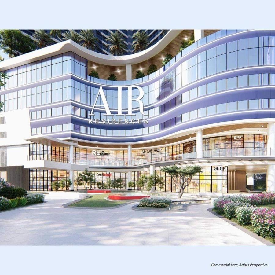 The exterior of the Air Residences building is displayed, featuring a modern architectural design with curvilinear glass facades. Lush greenery and flowering plants are visible in the landscaped area, enhancing the inviting appearance of the commercial space below.