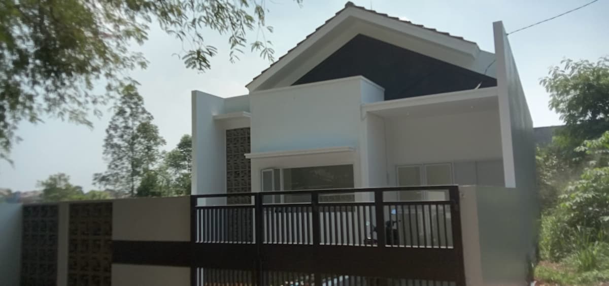 A modern, single-story house with a sleek design features a distinctive black roof and large windows that invite natural light. The property is enclosed by a simple fence, surrounded by lush greenery, adding to its serene ambiance.