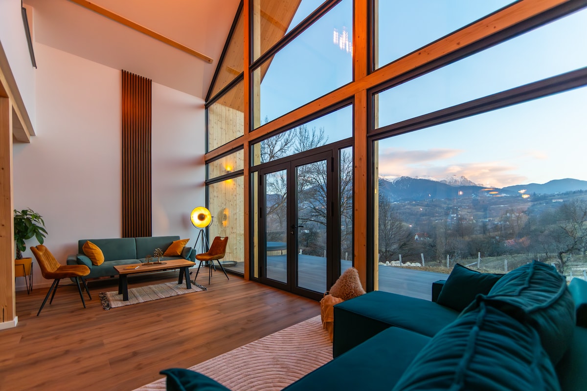 The spacious living area features large glass windows offering stunning views of the surrounding mountains. A cozy seating arrangement is set up with green sofas and armchairs. A stylish coffee table sits at the center, while warm lighting adds to the inviting ambiance.