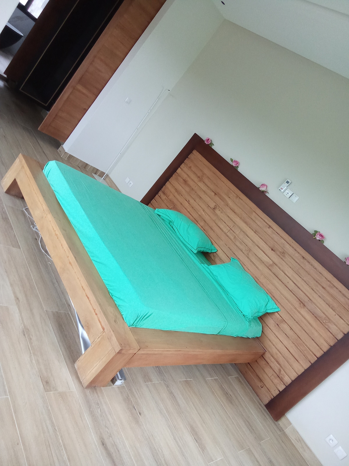 A spacious sleeping area features a wooden bed frame with a green bedding set. The room is designed with a natural wood accent wall and light-colored wooden flooring, providing a serene and open atmosphere.