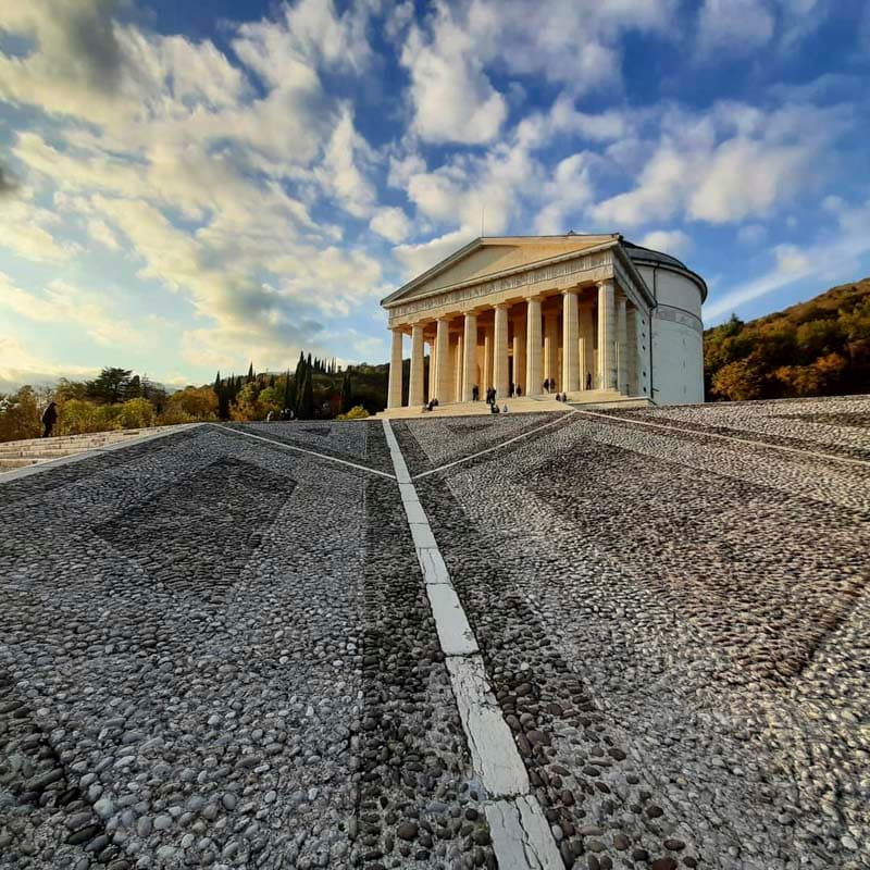 A grand building with tall columns occupies the foreground, complemented by a striking stone pathway leading up to it. The sky is partly cloudy, allowing soft light to illuminate the surrounding landscape, which features trees on the hillside.