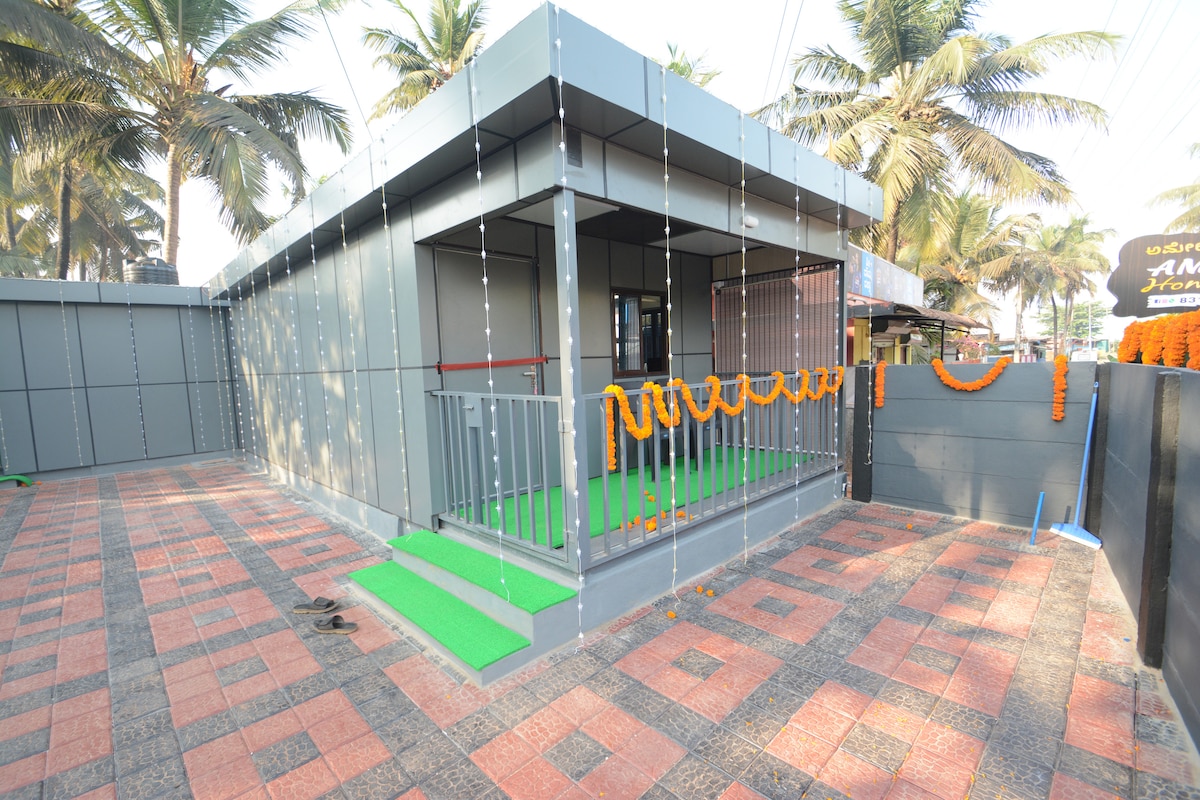 The exterior features a modern building surrounded by palm trees, with a spacious patio paved in patterned tiles. Decorative marigold garlands hang along the entrance, adding a festive touch. A green lawn mat leads up to the entrance, enhancing the welcoming appearance of the space.