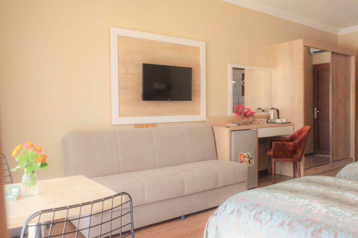 A modern room features a soft grey sofa and a small dining table with a vase of flowers. A flat-screen TV is mounted on the wall above a light wood console. A cozy armchair adds a pop of color to the inviting space.