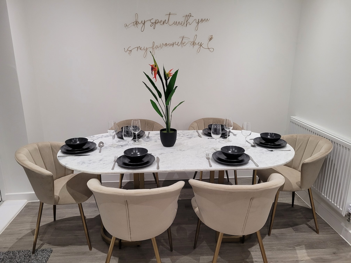 A circular marble dining table is set for six, surrounded by beige upholstered chairs. A small potted plant with vibrant flowers sits in the center, complementing the elegant table setting, which includes black plates and glassware.