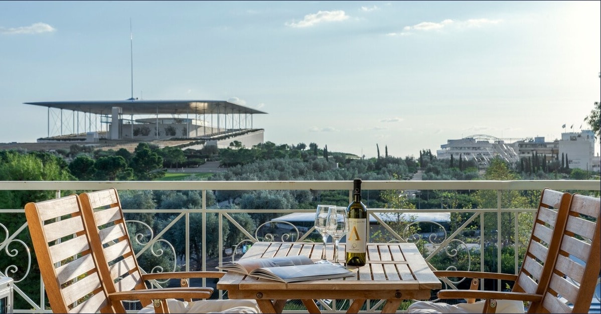 A balcony table is set with a bottle of wine and glasses, overlooking the lush greenery and the modern architectural silhouette of the Stavros Niarchos Foundation Cultural Centre in the distance, enhancing the tranquil ambiance.