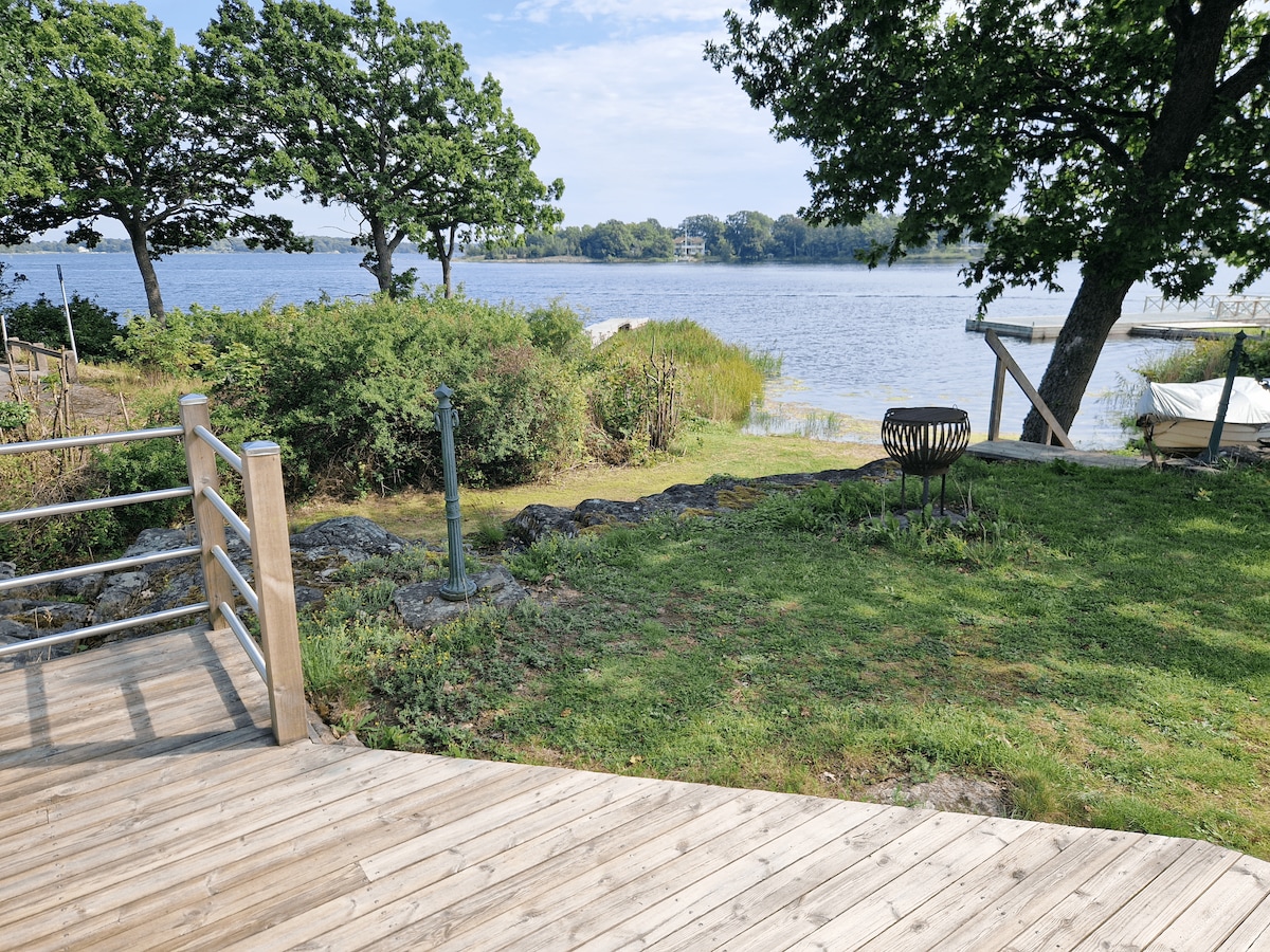 A serene view of the waterfront is captured, featuring lush greenery and a wooden deck leading towards the water. A fire pit is positioned on the grassy area, with gentle waves visible in the distance under a clear sky.