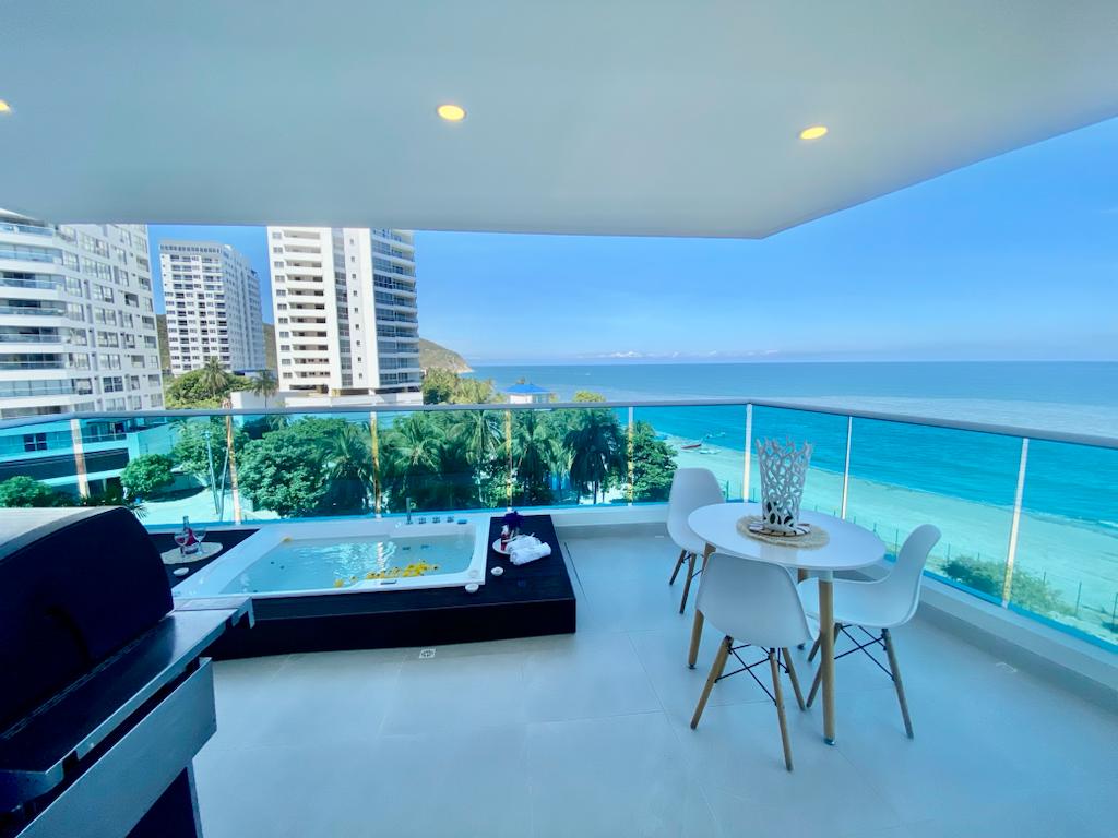 A modern outdoor balcony features a dining area with a round table and white chairs overlooking the ocean. A hot tub is situated nearby. The tranquil beach and palm trees can be seen in the background, with tall buildings framing the scene.