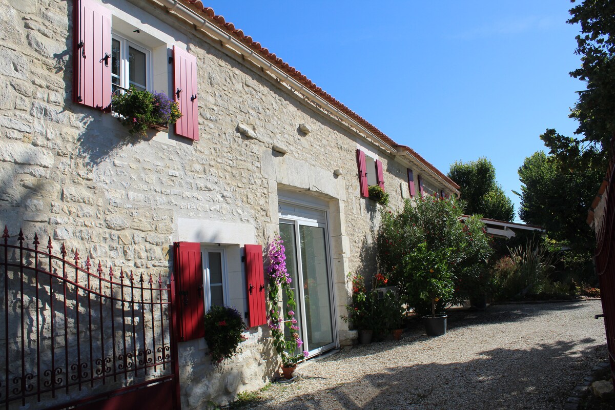 A charming stone exterior is featured, accented by vibrant red shutters and window boxes filled with flowers. A gravel pathway leads to the entrance, flanked by greenery and small trees, creating a serene and welcoming atmosphere.