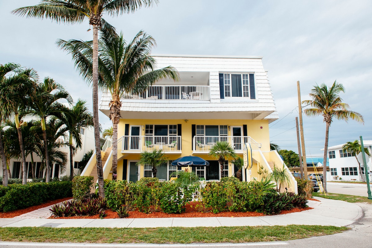 A tropical building features two levels with a bright yellow façade and white trim. Lush palm trees and manicured landscaping surround the entrance, which is adorned with an umbrella. The covered balconies on the upper floor overlook the inviting outdoor space.