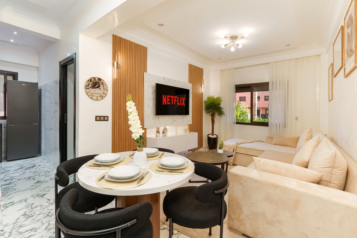 A modern living space features a 60-inch smart TV displaying Netflix on a sleek wall. A round dining table with four black chairs is set for meals. A comfortable, light-colored sofa bed complements the decor, while large windows allow natural light to brighten the area.