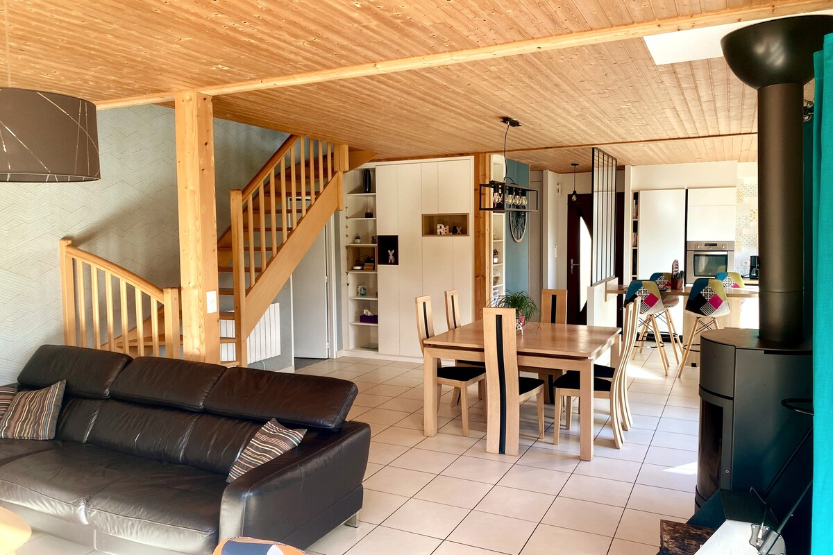 An open living area features wooden beams and a modern design, showcasing a comfortable leather sofa and a dining table with six chairs. A staircase leads to an upper level, while natural light illuminates the space from large windows.