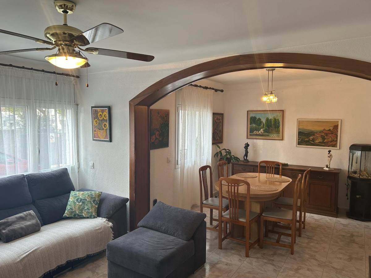 A spacious living area is visible, featuring a comfortable sofa with an accent pillow and a plush armchair. A dining table surrounded by wooden chairs is positioned next to a sideboard. Natural light filters through sheer curtains, illuminating the art on the walls.