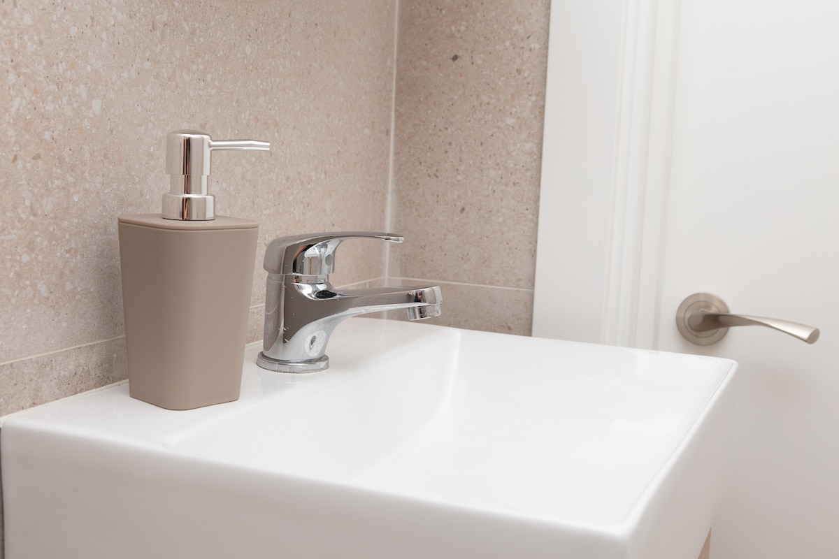 A modern bathroom sink setup is presented, featuring a sleek white basin and a contemporary faucet. A beige soap dispenser is positioned beside the faucet, accentuating the clean lines of the space. The wall displays a neutral, textured finish, adding subtle depth.