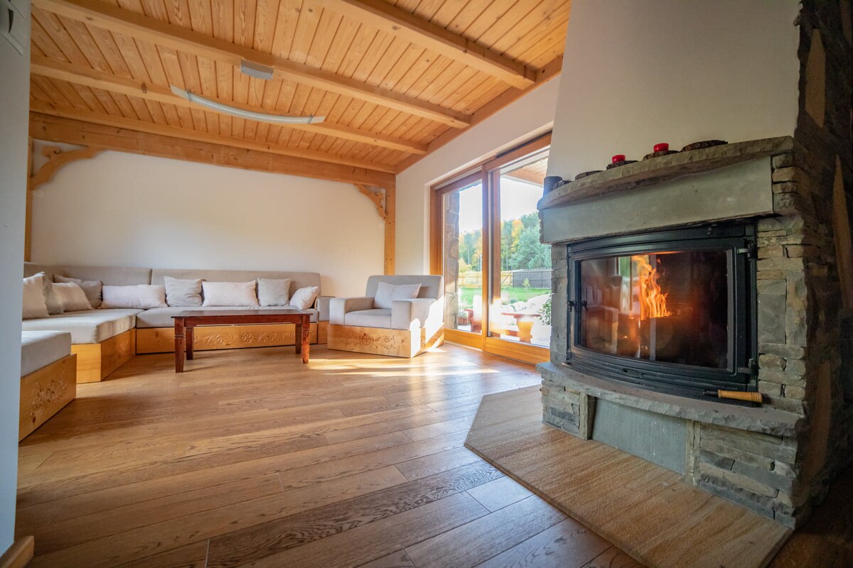 A cozy living area features a stone fireplace that provides warmth and a soft glow. Natural light fills the space through large windows, which offer views of the outdoors. A comfortable sectional sofa and a wooden coffee table complete the inviting setting.