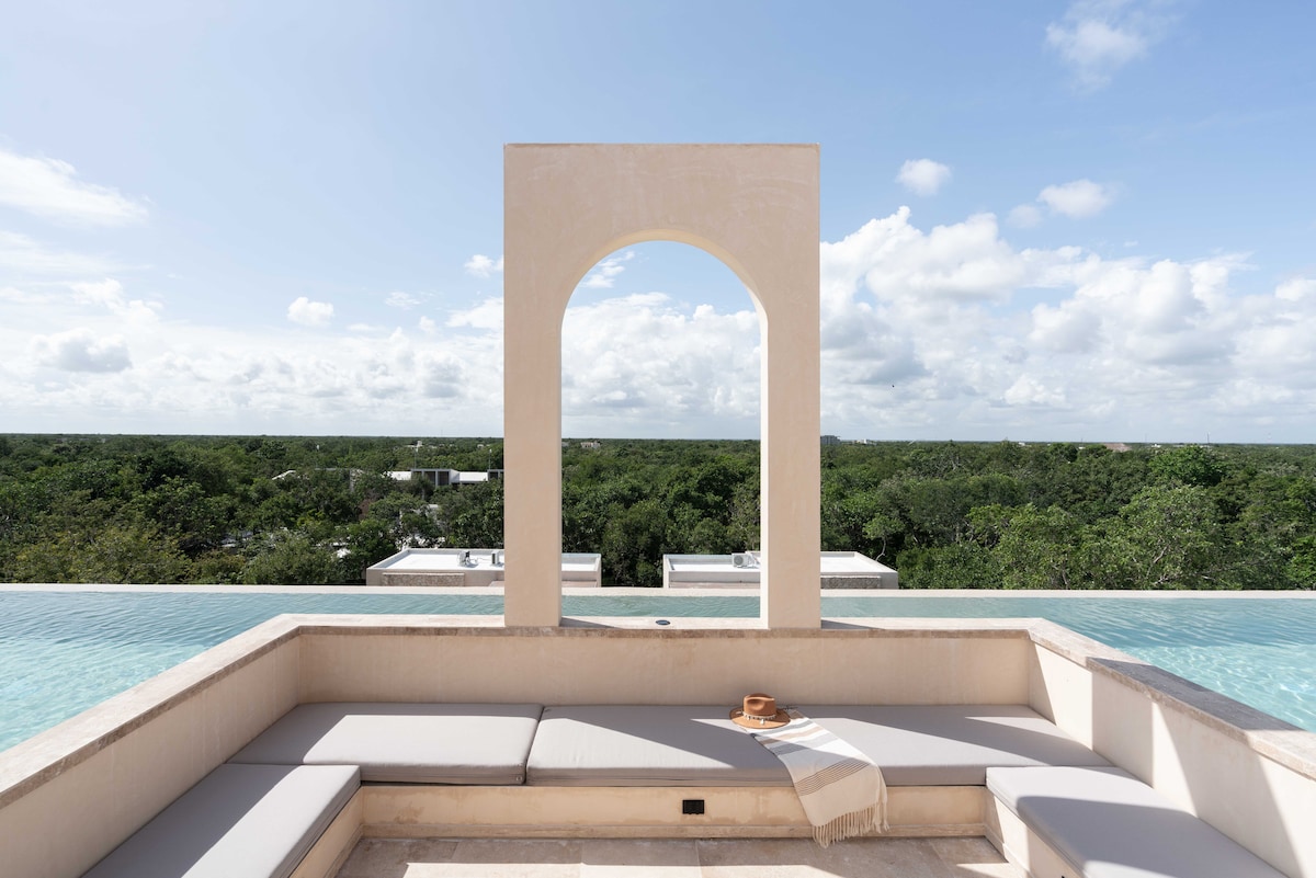 A lounge area is framed by a striking arch, overlooking a lush green landscape. Soft seating with light grey cushions provides a comfortable spot for relaxation. The infinity pool reflects the sky, creating a serene connection with nature.