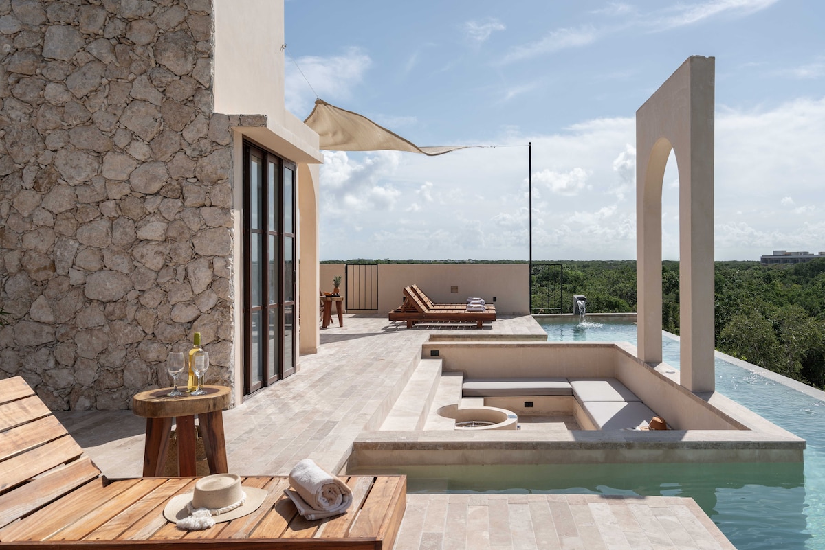 A tranquil outdoor area features a private pool with an infinity edge overlooking the lush jungle. Lounge chairs and a small table are positioned beside the pool, inviting relaxation. An architectural arch and soft fabric canopy add visual interest, while textured stone walls create a natural ambiance.