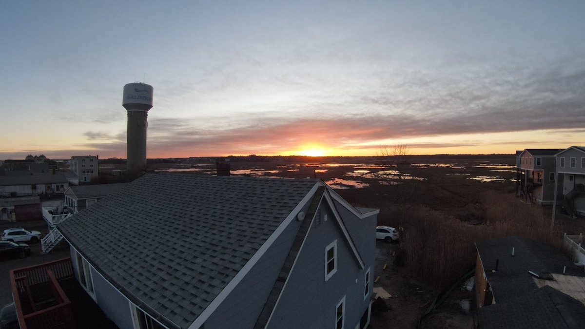 A panoramic view captures a serene sunrise over wetlands, with vibrant hues reflected in the calm water. Nearby buildings and structures are visible, creating a sense of community. A water tower stands prominently, blending into the tranquil landscape.