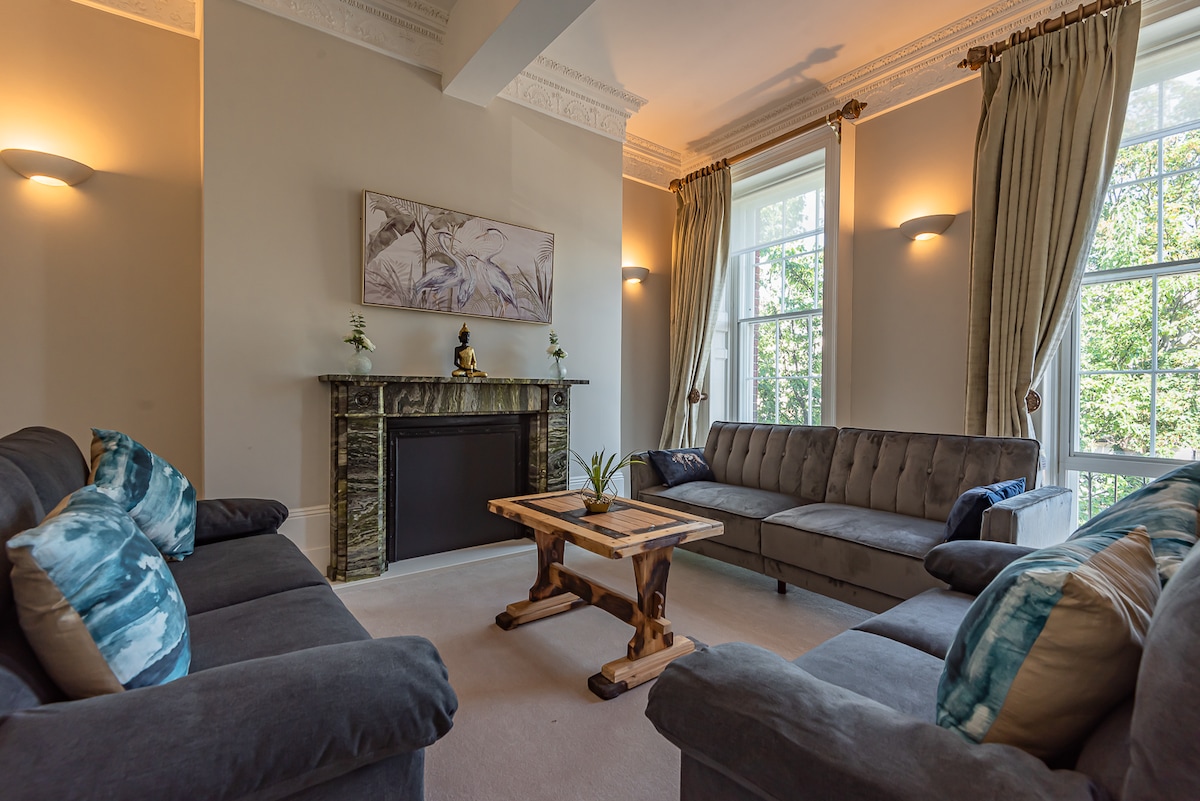 A spacious living area features two plush sofas with decorative cushions, complemented by a wooden coffee table. Sunlight filters through large windows dressed in light curtains, illuminating the elegant design elements, including a classic mantelpiece adorned with decorative items.
