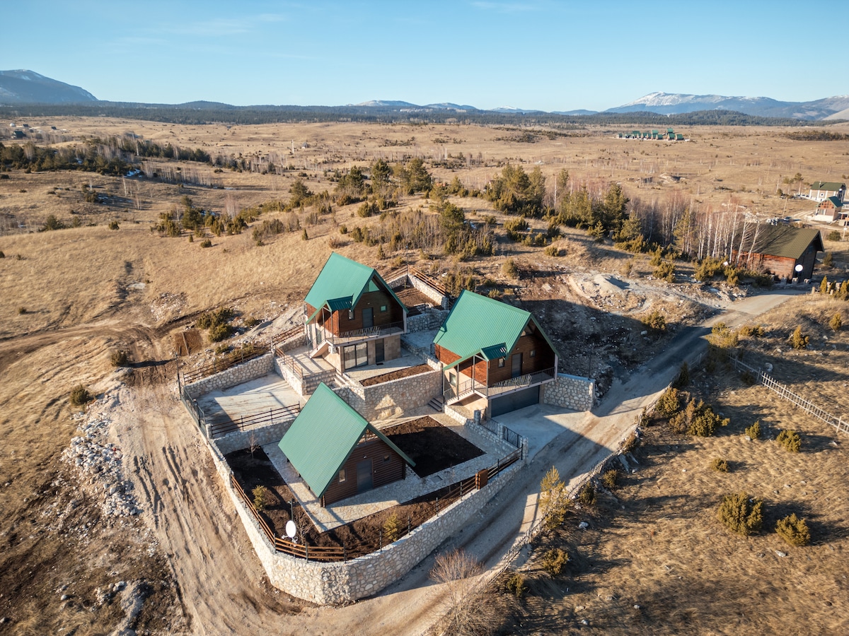 Aerial view of a charming getaway featuring three wooden cottages with green roofs, set against a sprawling landscape of open fields and distant mountains. The cottages are arranged on a stone base with pathways leading to each structure, surrounded by natural vegetation.
